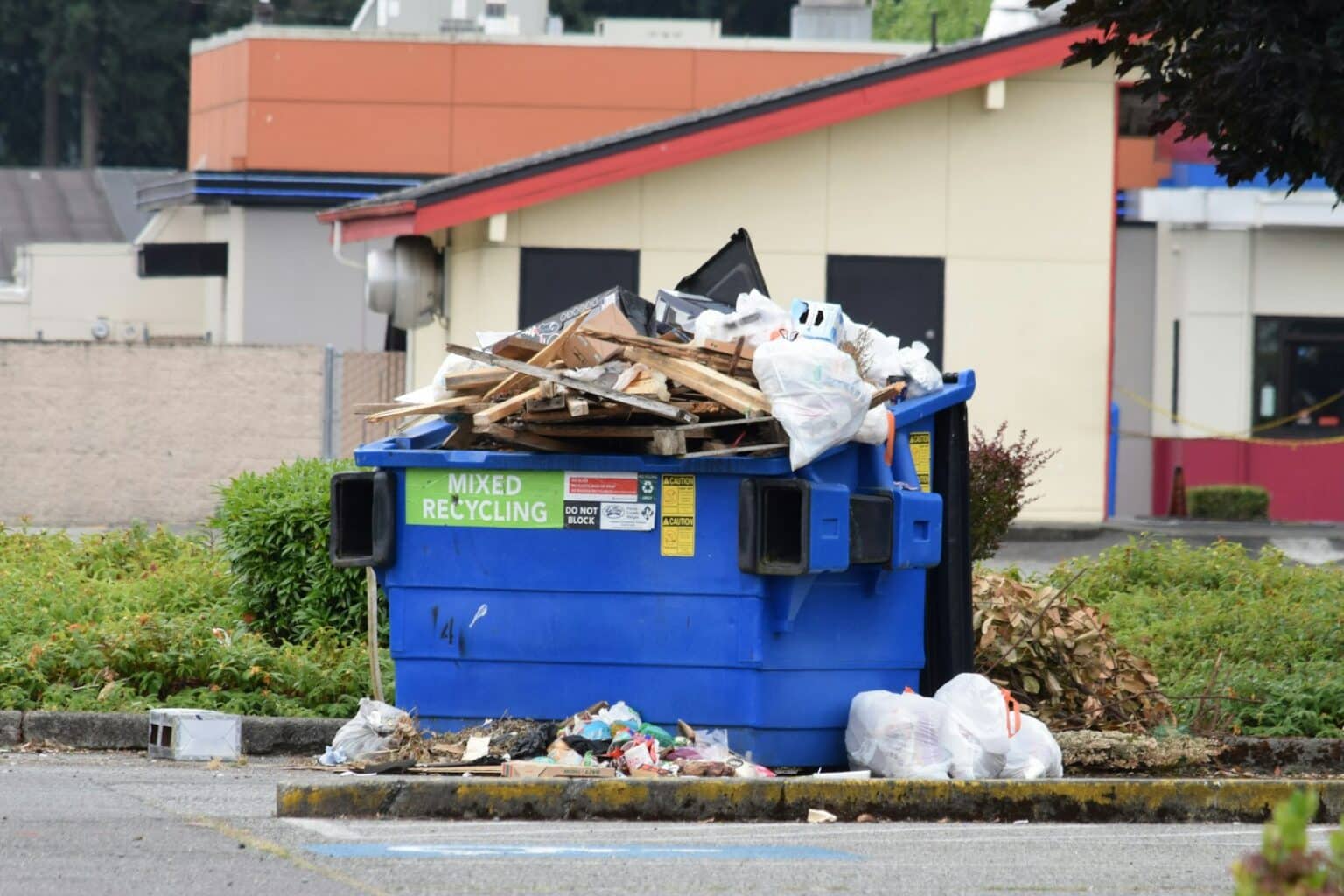 Blue mixed-recycling dumpster overflowing with wood scraps, cardboard, and bags