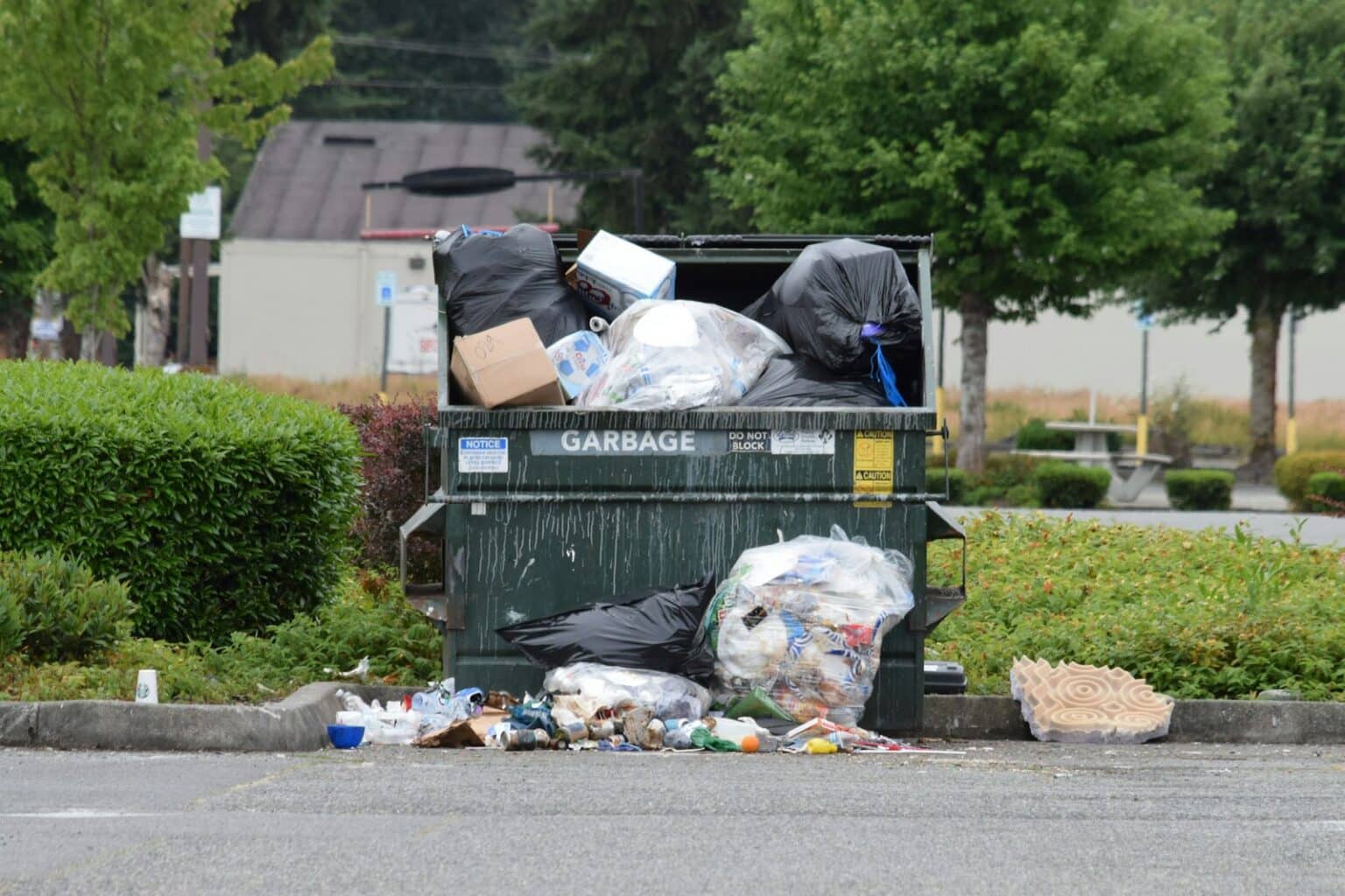 Large green metal dumpster overflowing with trash bags on an asphalt lot